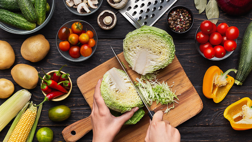 cutting board with hands chopping a cabbage with other fresh vegetables laid out, tomatoes, cucumbers, peppers, corn, potatoes